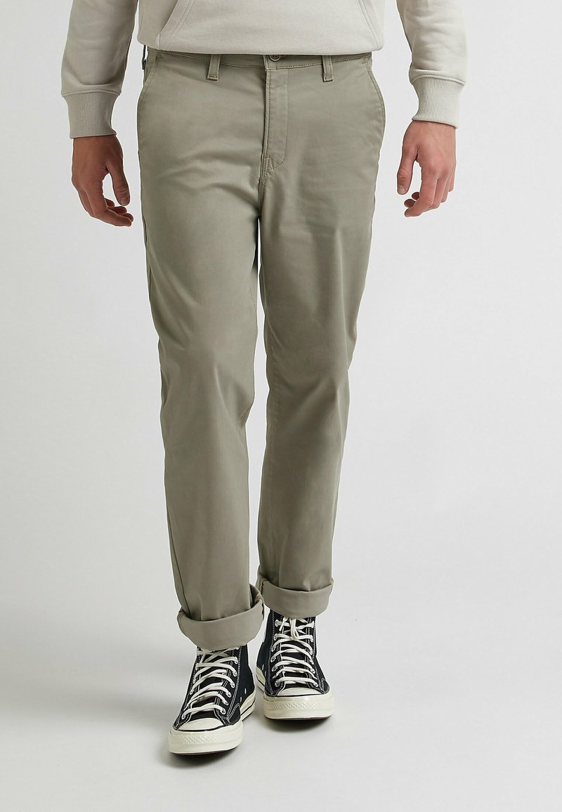 Homme portant un pantalon beige avec les revers roulés, des baskets montantes noires à lacets blancs, et un sweat-shirt de couleur claire.