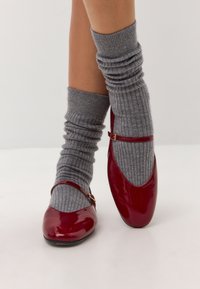 Scarpe in vernice rossa con punta arrotondata e cinturino a fibbia, abbinate a scaldamuscoli in maglia grigia a coste.