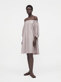 Femme debout portant une robe rose ample, épaules dénudées, longueur genou, avec des manches bouffantes et des sandales plates marron sur un fond uni.