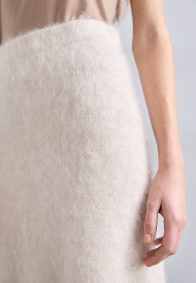 Beige, blødt tekstureret nederdel lavet af uld med en fuzzy overflade. Nærbillede af stofdetaljer, hånd hvilende ved siden af nederdelen.