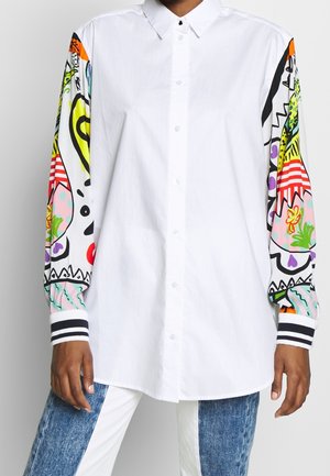 Weißes Hemd mit Knöpfen und mehrfarbiger abstrakter Kunst auf den Ärmeln, schwarz-weiß gestreifte Manschetten, kombiniert mit blau-weißen Jeans.