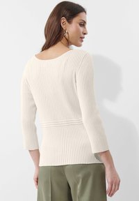 Beige gerippter Strickpullover mit einem tiefen Rückenausschnitt und drei Viertelärmeln, der über strukturierte Streifen und einen taillierten Bund verfügt.