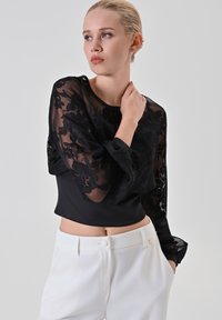 Zwarte blouse met doorzichtige, bloemenkant lange mouwen, een aansluitend lijfje en een kort ontwerp. Gepaard met witte hoge taillebroeken.