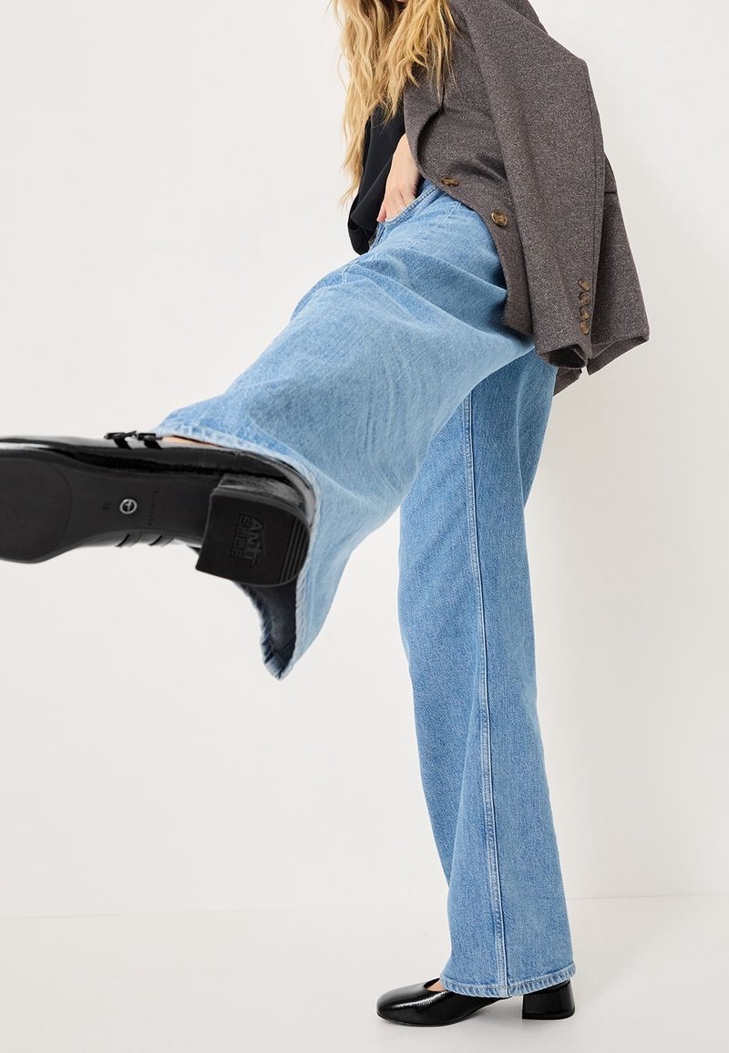 Personne portant un jean bleu large, des chaussures noires et un blazer marron, levant une jambe vers l'avant sur un fond blanc uni.