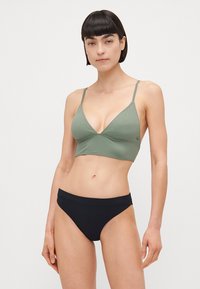 Zelený trojuholníkový bralet s tenkými ramienkami a bezšvovým dizajnom, kombinovaný s čiernymi bikinkami, ktoré majú hladký pás a minimálne zakrytie.
