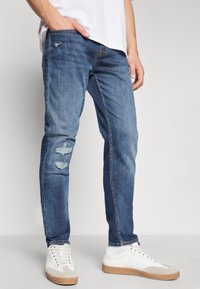 Jean en denim bleu clair avec une coupe slim, présentant des déchirures usées au niveau des genoux, associés à des baskets blanches. Texture lisse, design classique.