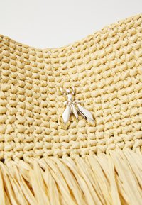 Sac en paille tissée de couleur jaune clair, avec un détail de broche en métal en forme d'insecte et une frange le long du bord inférieur.