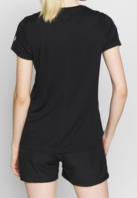 Frau mit blonden Haaren in einem schwarzen kurzärmeligen Shirt und schwarzen Shorts, die von einem schlichten hellen Hintergrund abgewandt ist.