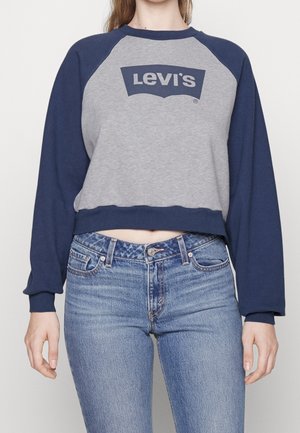 Kobieta ubrana w niebiesko-szarą bluza Levi’s z długim rękawem i wysokim stanem niebieskie dżinsy, stojąca z rękami swobodnie opuszczonymi wzdłuż ciała.