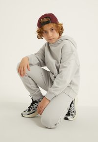 Pelēks kapucis un saskaņotas treniņbikses, abas izgatavotas no mīkstas materiāla. Melni un sudraba sporta apavi ar teksturētām detaļām. Tumši sarkana cepure.