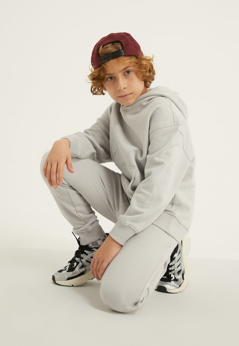 Pelēks kapucis un saskaņotas treniņbikses, abas izgatavotas no mīkstas materiāla. Melni un sudraba sporta apavi ar teksturētām detaļām. Tumši sarkana cepure.