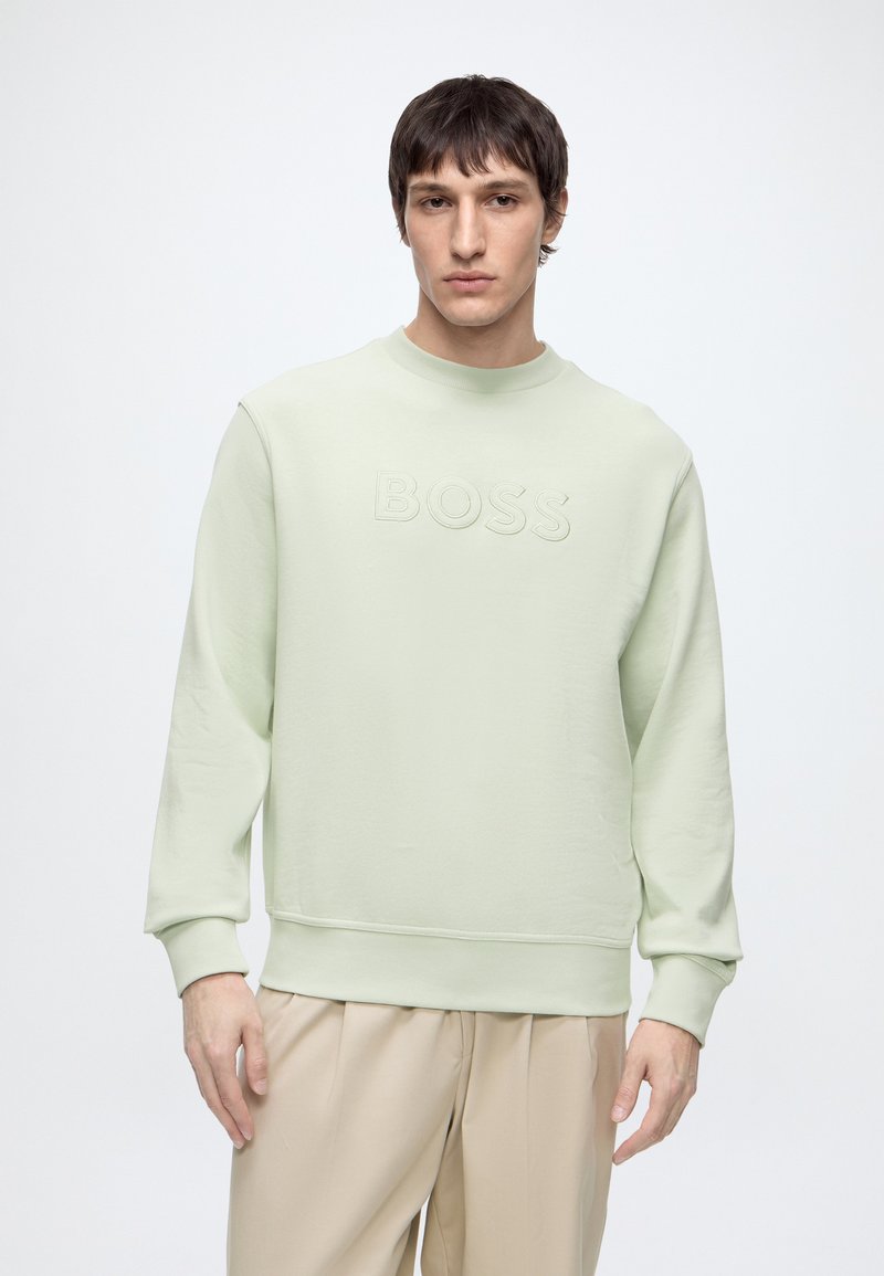 Jeune homme portant un sweat-shirt vert clair avec le texte en relief « BOSS » et un pantalon beige, debout sur un fond blanc uni.