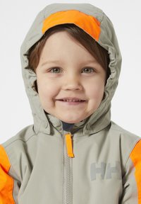 Lichtgrijze jas met oranje accenten. Voorzien van een rits aan de voorkant, capuchon en logo op de borst. Gemaakt van glad, waterdicht materiaal.