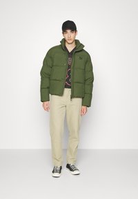 Giacca a vento verde con colletto alto, chiusura con zip e polsini elasticizzati. Indossata sopra una maglietta nera con grafica e abbinata a pantaloni beige e sneaker nere.