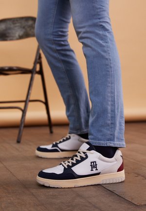 Benen in lichtblauwe spijkerbroek en witte sneakers met zwarte details, staand naast een zwarte klapstoel op een houten vloer.