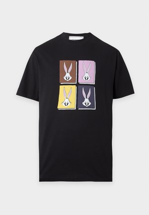 Mand iført sort t-shirt med farverige kaninillustrationer, brede mørkeblå jeans og sorte sko, der står mod en hvid baggrund.