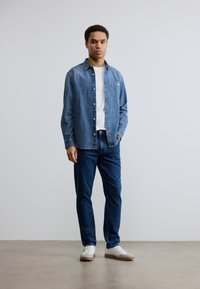 Πουκάμισο από τζιν σε ανοιχτό μπλε χρώμα με κλείσιμο με κουμπιά, τσέπη στο στήθος· φορεμένο πάνω από λευκό μπλουζάκι και συνδυασμένο με σκούρο μπλε τζιν και λευκά αθλητικά παπούτσια.