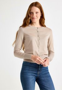 Cardigan beige realizzato in morbido tessuto a maglia, con scollatura rotonda e chiusura frontale con bottoni. Abbinato a jeans blu scuro.