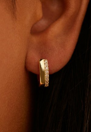 Gouden creool met een rij kleine heldere edelstenen, gedragen aan de oorlel van een persoon met een middel-donkere huid.