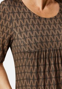 Braun gemusterte Bluse mit geometrischem Design in Schwarz, runder Ausschnitt, kurze Ärmel und Raffung in der Taille.