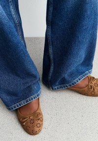 Bruine geperforeerde leren ballet flats met een strik; wijde donkere blauwe denim jeans; lichte gestructureerde vloer achtergrond.