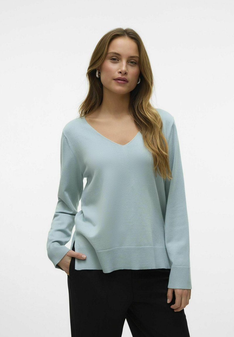 Maglione azzurro chiaro a maniche lunghe con scollo a V, texture morbida, fessure laterali e polsini a coste. Indossato con pantaloni neri, design semplice, senza motivi.