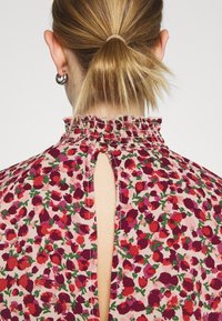Blommig blus i rosa tyg med röda och gröna mönster, volangkrage och nyckelhål i ryggen.