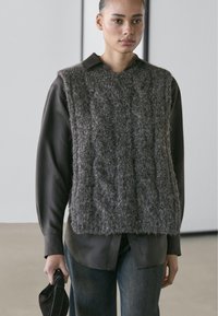 Gray, textured knit vest with cable pattern over a dark shirt. Model holds a dark cap. Neutral background emphasizes the outfit’s layering.