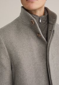 Sivý kabát z vlny s vysokým golierom, vybavený drevenými gombíkmi a zipsom vo vnútornej vrstve. Hladká textura a prispôsobený strih.