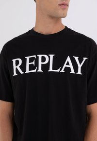 Camiseta de algodón negra con el texto en blanco "REPLAY" en el pecho. Ajuste regular con mangas cortas y cuello redondo.