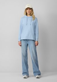 Felpa azzurra con cappuccio e tasca frontale, abbinata a jeans azzurri a gamba larga. Sneakers bianche e un cappello beige completano il look.