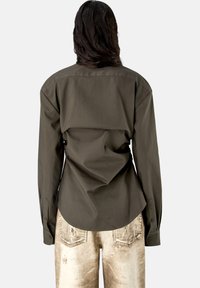 Persona con cabello largo y oscuro que lleva una camisa de manga larga de color oliva oscuro y vaqueros claros desgastados, vista desde atrás sobre un fondo liso.