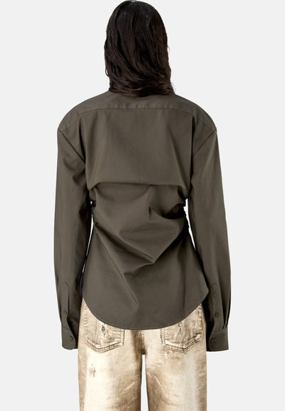 Persona con cabello largo y oscuro que lleva una camisa de manga larga de color oliva oscuro y vaqueros claros desgastados, vista desde atrás sobre un fondo liso.