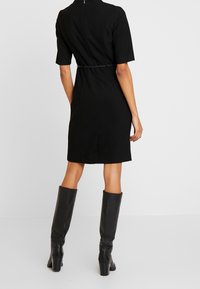 Schwarzes, knielanges Kleid mit kurzen Ärmeln, tailliertem Bund und Reißverschluss hinten. Kombiniert mit knielangen schwarzen Lederstiefeln. Schlichte Textur.
