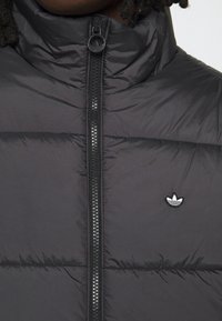 Černá prošívaná péřová bunda s vysokým límcem, zapínáním na zip a horizontálním stehováním. Výrazné bílo-černé logo na hrudi.