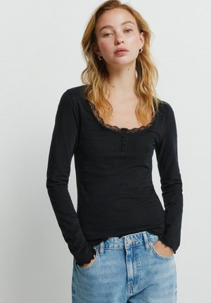 Blondynka z rozpuszczonymi włosami, ubrana w czarną bluzkę z długim rękawem i koronkowym wykończeniem oraz jasnoniebieskie dżinsy, ręce w kieszeniach, neutralne tło.