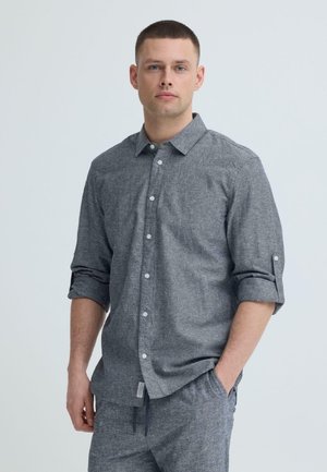 Man met kort haar, gekleed in een grijs overhemd met opgerolde mouwen en bijpassende grijze broek, staand tegen een effen lichte achtergrond.