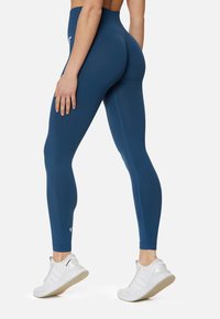 Leggings blu a vita alta realizzati in un tessuto liscio e elastico, con un logo discreto alla caviglia, abbinati a scarpe da ginnastica bianche.