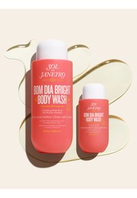 Dos botes de gel de ducha corporal Sol de Janeiro Bom Dia Bright de color coral, uno grande y uno pequeño, con salpicaduras de gel transparente detrás de ellos.