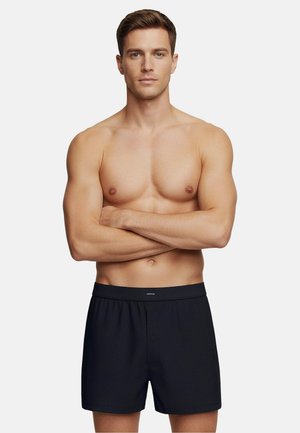 Černé bavlněné boxerky s knoflíkovým zapínáním a elastickým pasem, zobrazené na mužském modelu se zkříženýma rukama a odkrytým hrudníkem.