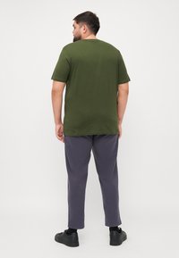 Olijfgroene t-shirt met korte mouwen en ronde hals, gecombineerd met grijze tapse broek en zwarte sneakers, gezien vanaf de achterkant.