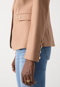 Beige sako se zlatým kováním, jednou náprsní kapsou a knoflíkovými manžetami, spárované se světle modrými džínami. Hladká textilie.