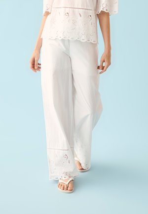 Mujer con pantalones anchos de encaje blanco con ojales y top a juego con chanclas, caminando sobre un fondo azul claro.