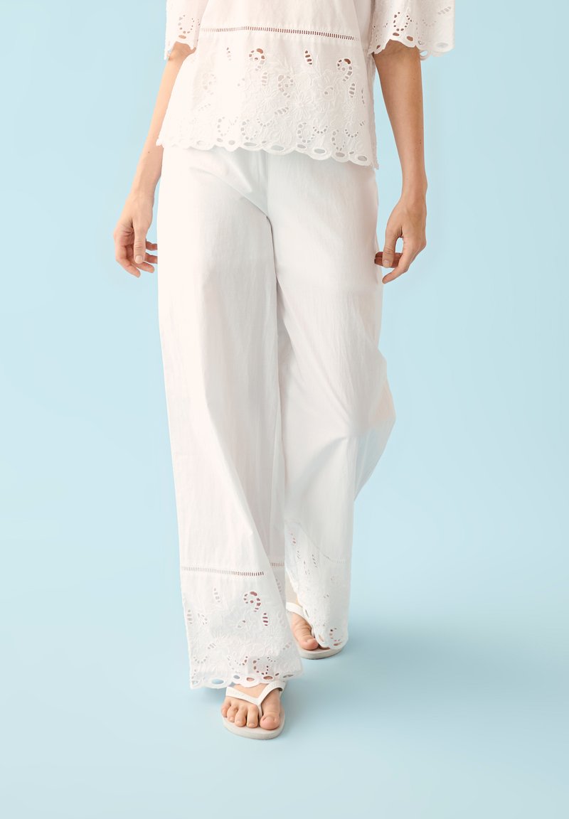 Femme portant un pantalon large en dentelle à œillets blanc et un haut assorti avec des tongs, marchant sur un fond bleu clair.
