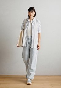 Camicia a righe blu e bianca, canotta bianca a costine, jeans larghi in lavaggio chiaro, sneakers grigie, che trasporta una grande borsa a tracolla beige.