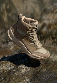 Bota de caminhada bege com uma mistura de tecido texturizado e sobreposições lisas, suporte alto para o tornozelo e sola robusta para tracção.