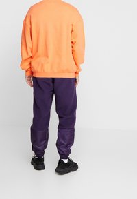 Pomarańczowa, oversize'owa bluza z prążkowanymi mankietami, zestawiona z fioletowymi dresami z polaru z błyszczącymi, fioletowymi panelami przy kostkach oraz czarnymi sneakersami.