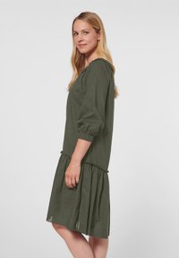 Olivenfarbiges Kleid mit lässiger Passform, Puffärmel und gerafftem Rüschensaum. Hergestellt aus weichem Stoff, mit einem runden Ausschnitt und strukturierten Details.