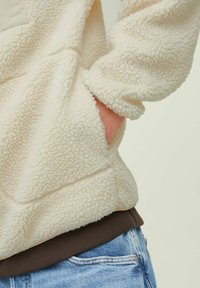 Persona che indossa una giacca in fleece color crema con le mani nelle tasche anteriori, sopra una maglietta scura e jeans blu.