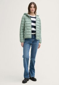 Lichtgroene pufferjack met capuchon, met horizontale stiksels, gedragen over een marineblauw en wit gestreept shirt, gecombineerd met blauwe flared jeans.